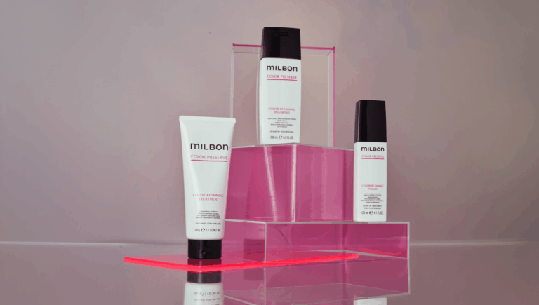 Milbon Color Preserve hair care trio on a pink acrylic display: a white tube on the left, a tall bottle in the center, and a smaller bottle on the right.