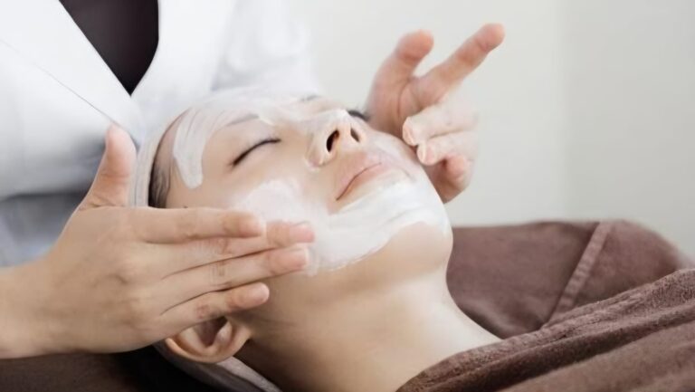 Esthetician applies a white facial mask to a relaxed client's face during a spa treatment, with gentle hand support on the chin and cheeks.
