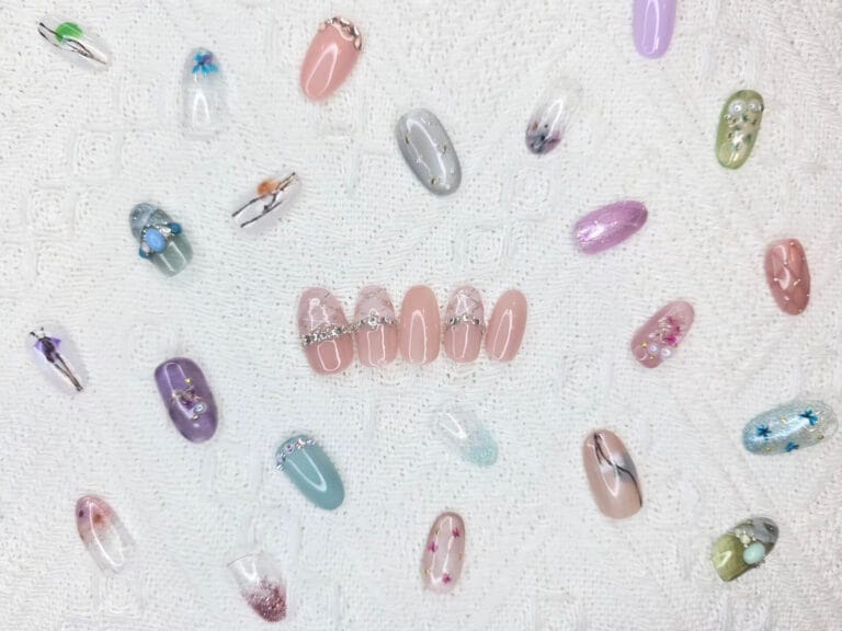 Circular arrangement of pastel acrylic nails with rhinestone accents on a white textured fabric background, viewed from above.