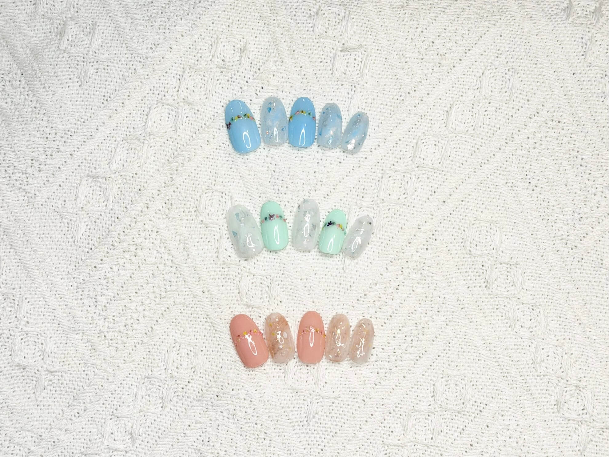 Pastel gel nail tips in blue, mint, and peach tones arranged in rows on a textured lace background, showcasing floral accents and glitter.