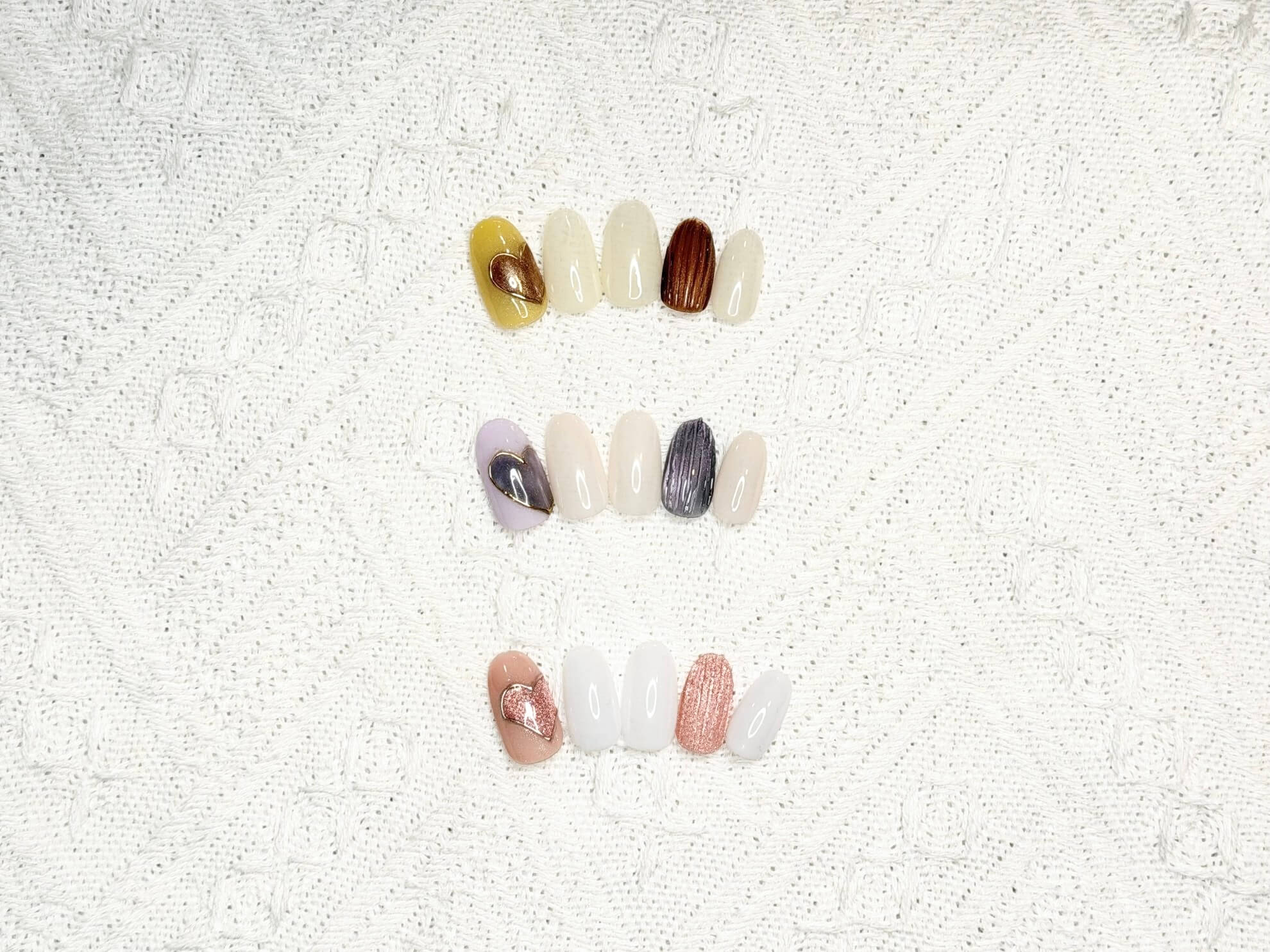 Row of artificial nail swatches on a white textured cloth, featuring nude, cream, and brown tones with metallic accents.
