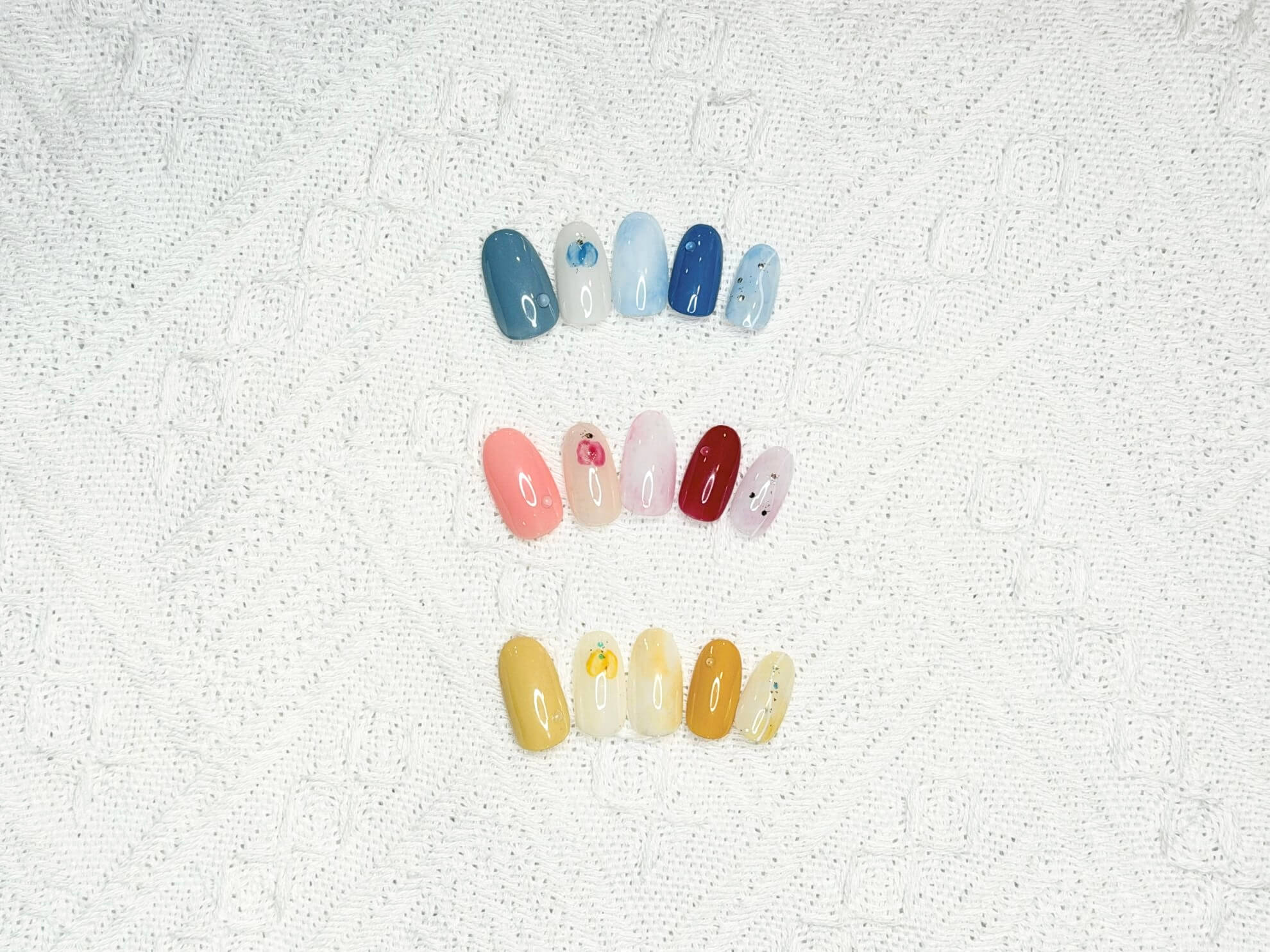 Three horizontal rows of artificial nails in different color families: blue tones on top, pink/peach and red in the middle, and yellow hues at the bottom, all with glossy finishes on a white textured towel background.