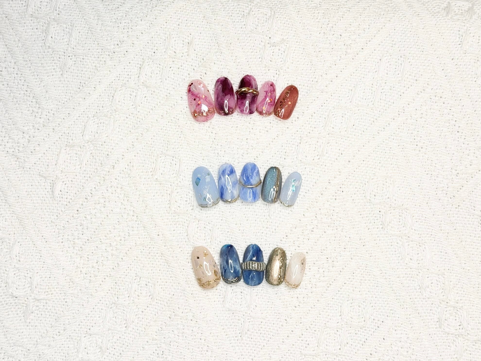 Rows of decorative nail tips arranged on a white textured background, with pink and purple marbled designs and gold flecks, showcasing nail art samples in two color families.