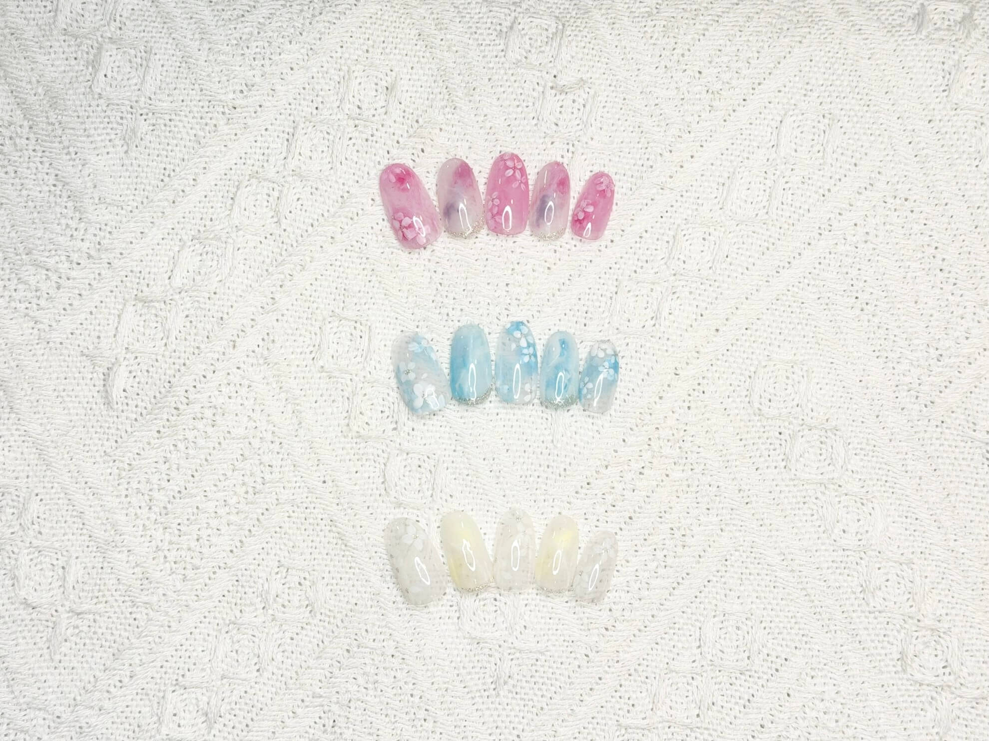 Row of pink and blue artificial nails with delicate white floral designs laid on a lace background