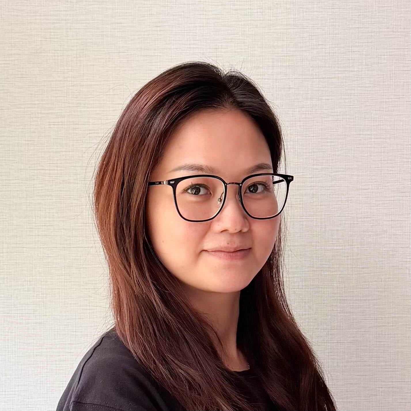 Portrait of a woman with long brown hair and black round glasses, facing slightly left against a light textured wall, smiling softly.