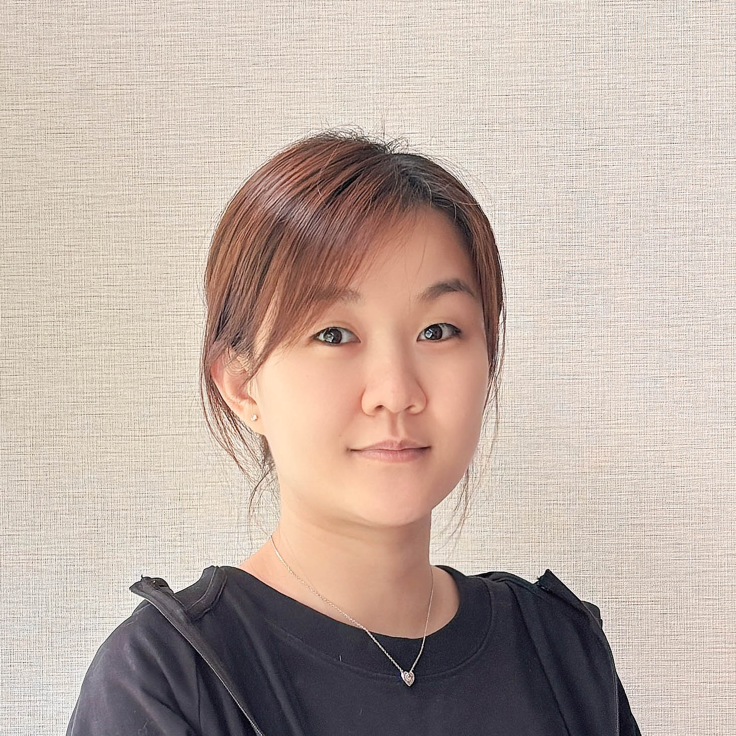 Portrait of a young woman with brown hair wearing a black top and a heart necklace, facing the camera with a neutral background.